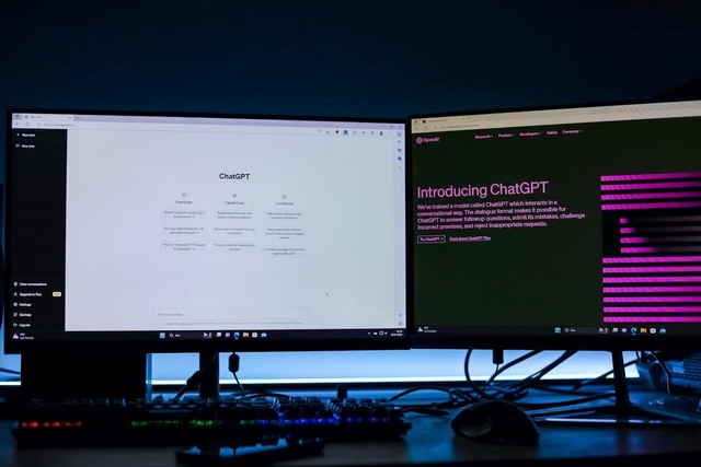 Dois monitores de computador lado a lado em uma mesa escura. A tela da esquerda exibe a interface de conversação do ChatGPT em modo claro. A tela da direita mostra o site oficial da OpenAI com a página de introdução ao ChatGPT sobre um fundo preto com grafismos em rosa